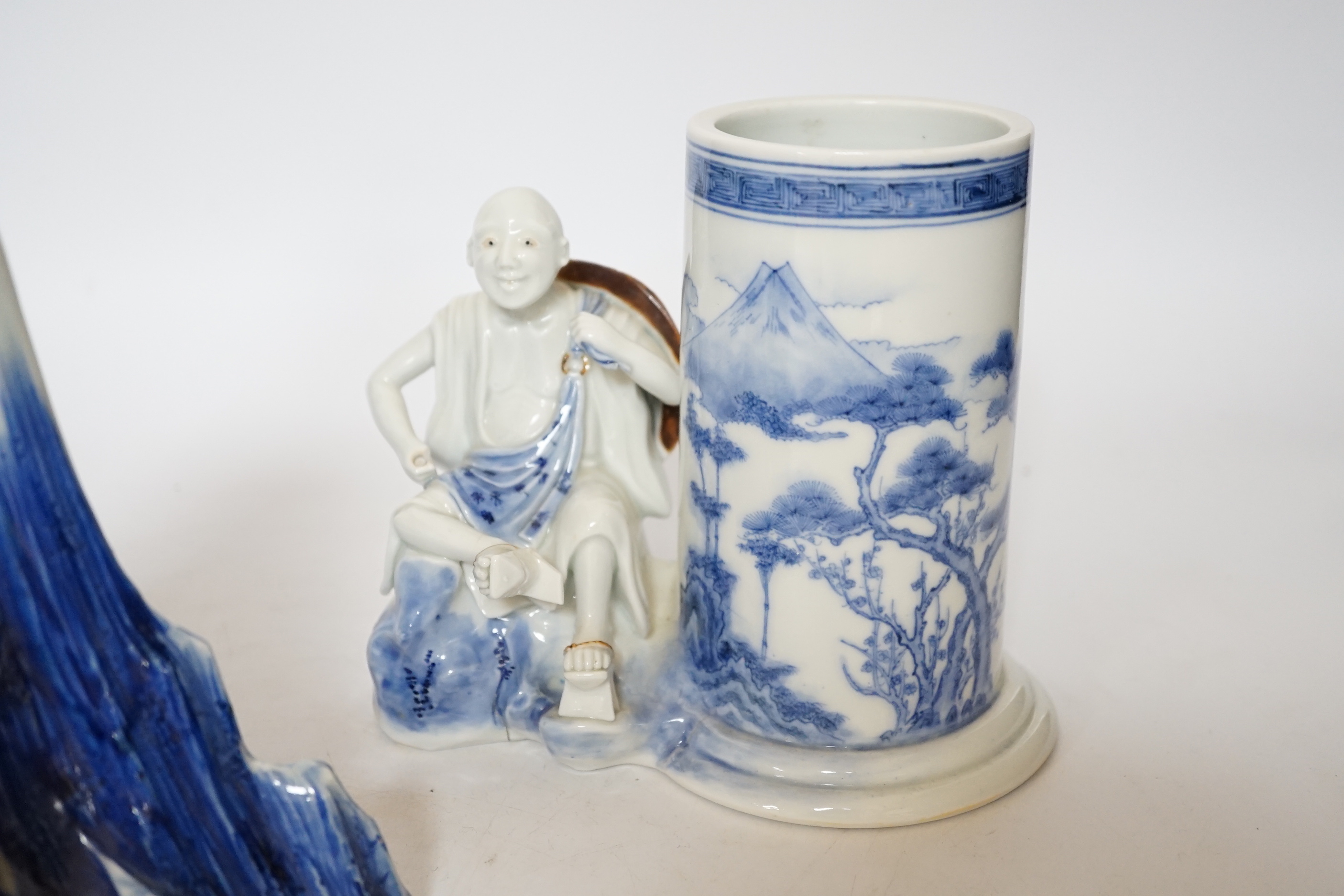 An early 19th century Japanese Hirado blue and white model of a monk seated by Mount Fuji, and a similar brush pot modelled with a fisherman seated by the cylindrical vessel, largest 17cm high (2)